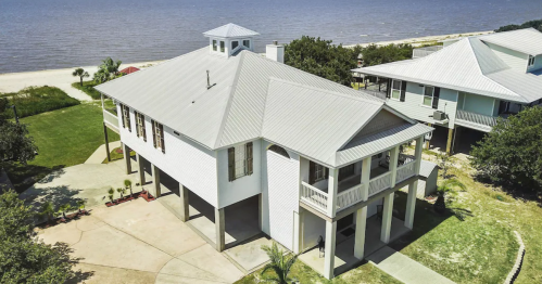 Aerial view of a modern beach house with a metal roof, surrounded by greenery and near a body of water.