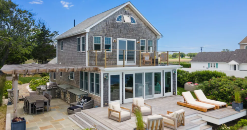 A modern two-story house with a deck, outdoor seating, and a landscaped yard under a clear blue sky.