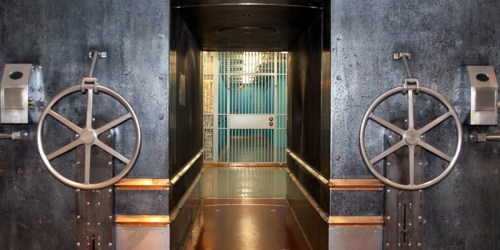 A view of a large, secure vault door with metal wheels, leading to a brightly lit area with barred gates.
