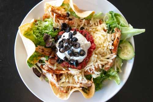 A colorful salad bowl with grilled chicken, cheese, olives, salsa, sour cream, and tortilla shells on a bed of greens.