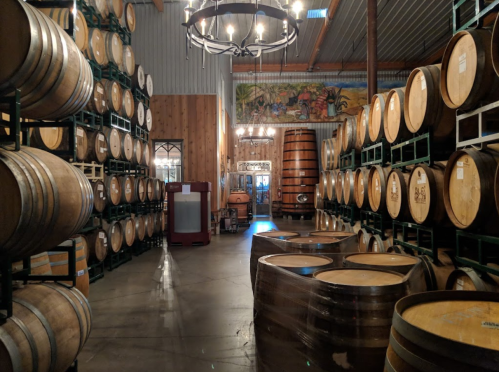 Interior of a winery with stacked wooden barrels, a chandelier, and a mural on the wall.