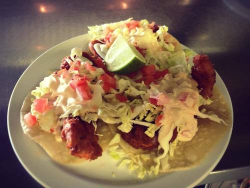 Three tacos topped with lettuce, tomatoes, and creamy sauce, served with a lime wedge on a white plate.