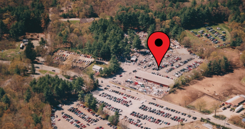 Aerial view of a large parking lot surrounded by trees, with a red location pin marking a specific area.