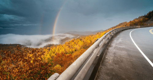 A winding road curves along a vibrant autumn landscape with colorful trees and a double rainbow in the misty mountains.