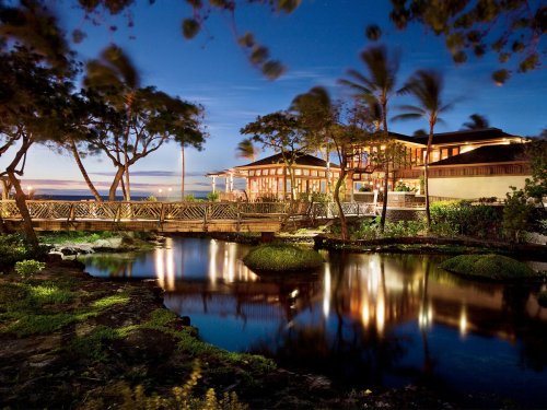 A serene waterfront scene at dusk, featuring a wooden bridge, palm trees, and a beautifully lit building reflecting in the water.