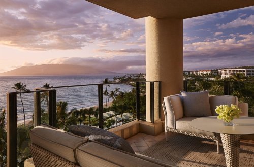 A serene balcony view overlooking the ocean, with palm trees and mountains in the distance during sunset.