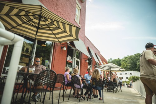A group of people sitting at outdoor tables under striped umbrellas outside a red brick building.