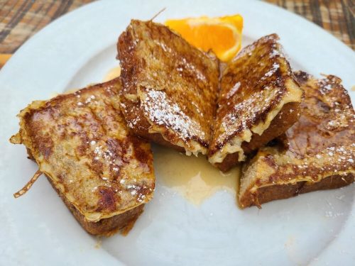Three pieces of French toast dusted with powdered sugar, served with a slice of orange and drizzled with syrup.