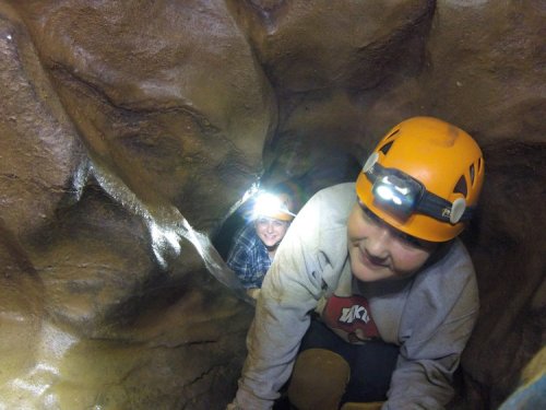 Two people in helmets with headlamps exploring a narrow cave passage, smiling and navigating through rocky terrain.