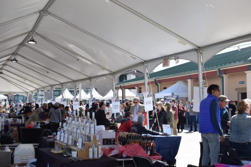A bustling outdoor market under a large tent, with vendors and visitors exploring various booths and products.