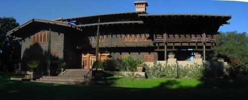 A large, two-story craftsman-style house with a wooden exterior, surrounded by greenery and a clear blue sky.