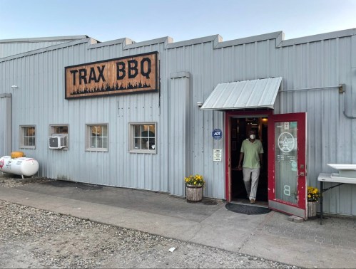 A person exits Trax BBQ, a metal building with a sign, surrounded by flowers and outdoor seating.