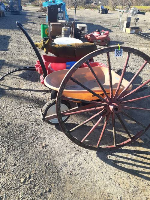 A red wheelbarrow with a large wheel, resting on gravel, surrounded by various tools and equipment in an outdoor setting.