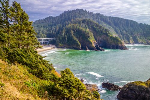 A scenic coastal view featuring a bridge, lush green hills, and waves crashing on the shore under a cloudy sky.