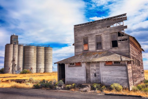 An abandoned wooden building next to large silos, set against a blue sky with scattered clouds and golden grass.