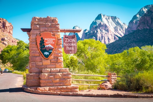 Zion National Park entrance sign with mountains and greenery in the background under a clear blue sky.