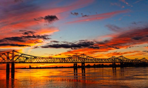 A sunset over a river, featuring a silhouetted bridge and vibrant clouds in shades of orange, purple, and blue.