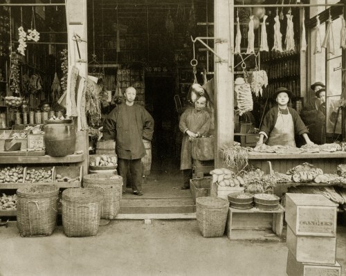 A historic market scene with four people outside a shop, surrounded by baskets of produce and hanging goods.