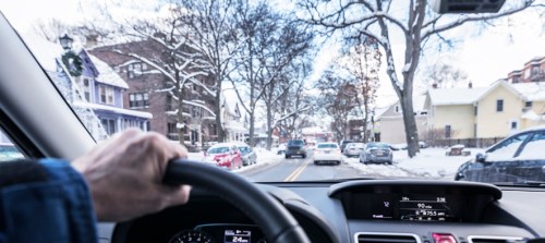 A driver's hand on the steering wheel, navigating a snowy street lined with parked cars and bare trees.