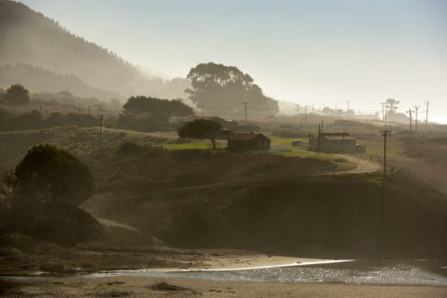 A misty landscape with rolling hills, a winding road, and distant houses under a soft, hazy sky.