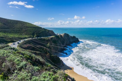 A scenic coastal view with a winding road, cliffs, and waves crashing against the shore under a blue sky.