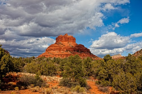 A red rock formation rises above green shrubs under a cloudy blue sky in a desert landscape.