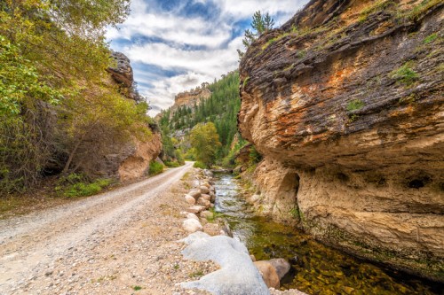 A scenic dirt path alongside a river, flanked by rocky cliffs and lush greenery under a cloudy sky.