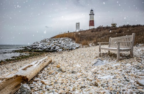 A snowy beach scene featuring a lighthouse, a bench, and rocky shoreline under a gray sky.