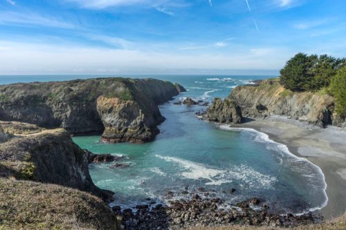A scenic coastal view featuring rocky cliffs, a calm bay, and gentle waves under a clear blue sky.
