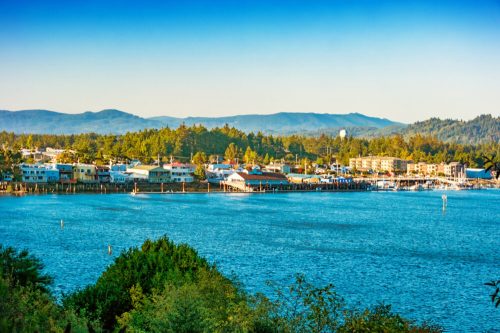 A scenic view of a coastal town with colorful buildings, a calm blue bay, and mountains in the background under a clear sky.
