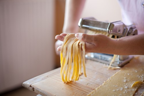 A person holds freshly made pasta strands above a wooden table, with a pasta machine in the background.