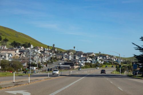A scenic road leads into a small coastal town, with houses on a hillside and clear blue skies above.
