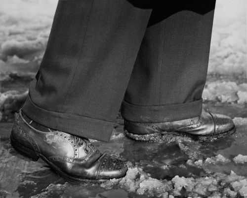 Close-up of polished leather shoes on a snowy surface, with tailored pants slightly rolled up at the cuffs.