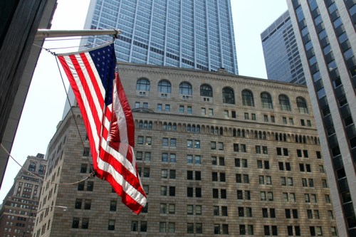 An American flag waves in front of tall buildings in a cityscape.