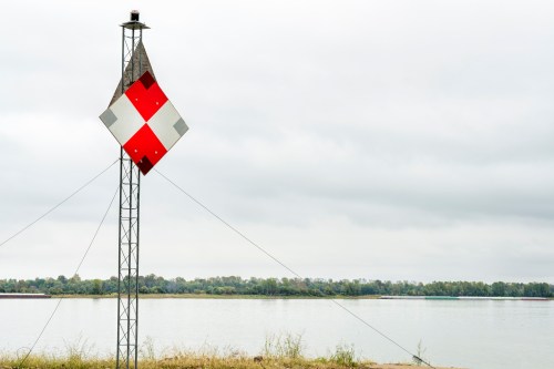 A red and white diamond-shaped sign on a pole by a calm river under a cloudy sky.