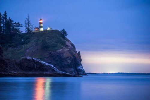 A lighthouse on a rocky cliff at dusk, with calm waters reflecting soft blue and orange hues.