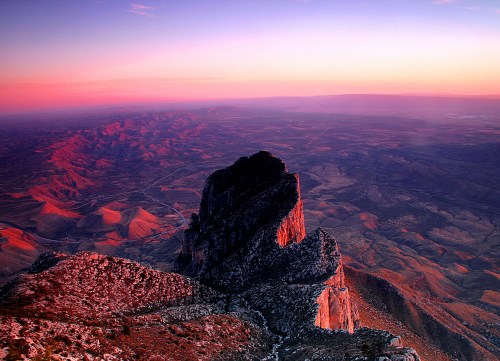 A stunning mountain peak at sunset, casting shadows over a vast, colorful landscape with rolling hills and valleys.