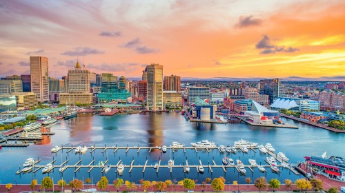 A vibrant sunset over a city skyline with boats docked in a marina and reflections on the water.