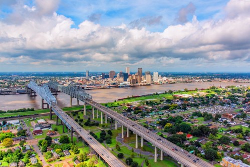 Aerial view of a city skyline with a river, bridge, and green landscape under a partly cloudy sky.