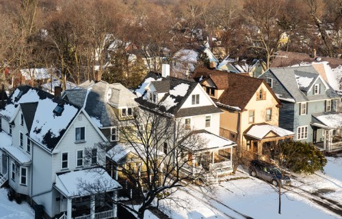 Aerial view of snow-covered houses in a residential area, with bare trees and a clear sky.