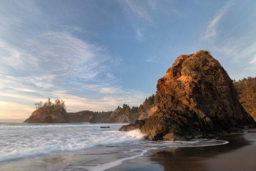 A rocky coastline at sunset, with waves gently crashing and a colorful sky filled with clouds.