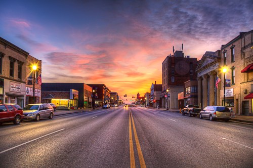 A quiet street at sunset, lined with buildings and parked cars, under a colorful sky.