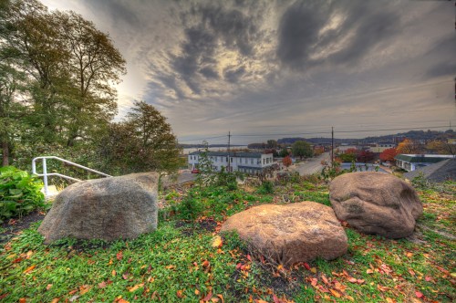 A scenic view from a hill featuring large rocks, trees, and a cloudy sky over a small town and river in the distance.