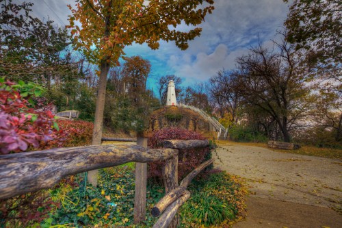A scenic pathway leads to a white lighthouse surrounded by colorful autumn foliage and trees under a cloudy sky.