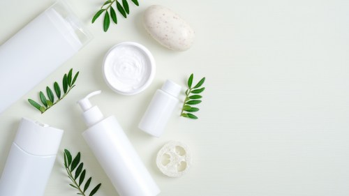 Flat lay of white skincare products, including bottles, a jar, and a soap bar, surrounded by green leaves.
