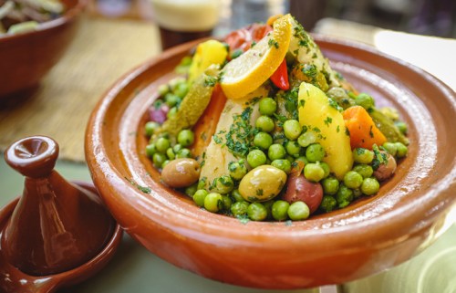 A colorful plate of couscous topped with vegetables and herbs, served in a traditional clay dish.