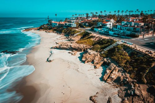 Aerial view of a coastal beach with rocky shores, palm trees, and houses along the shoreline under a clear blue sky.