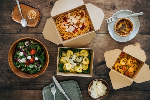 A variety of takeout meals on a wooden table, including pasta, salad, and a baked dish, with utensils nearby.
