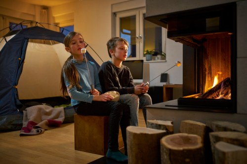Two children sitting by a fireplace, roasting marshmallows, with a tent set up in the background. Cozy indoor scene.