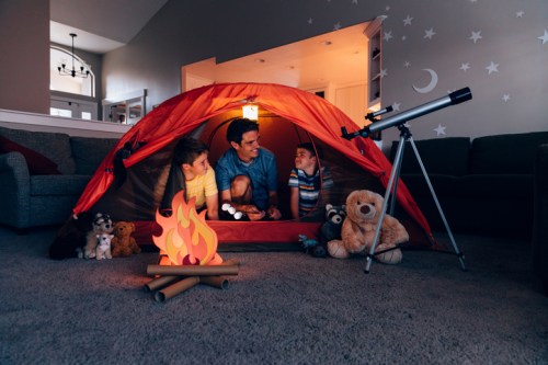 A man and two children sit inside a tent, surrounded by stuffed animals and a toy campfire, with a telescope nearby.